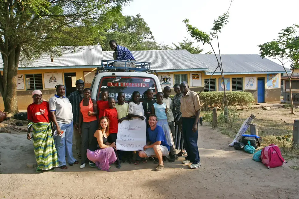 Todd, Christina, students and teachers in front of a van at a Ugandan school, holding a hand-lettered sign that reads Sean Lawson Young Travelers Foundation