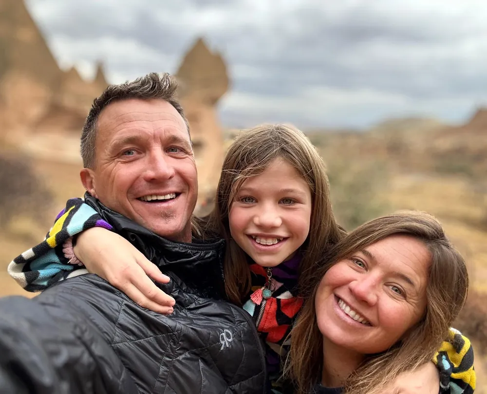 Todd Lawson and Christina Tottle smiling with their daughter on a trip, with rock formations behind them